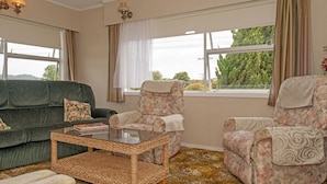 A cozy living room with a green sofa, a floral armchair, a wicker coffee table, and a view of greenery through large windows. - Holiday Haven - Whitianga Bach (Whitianga)
