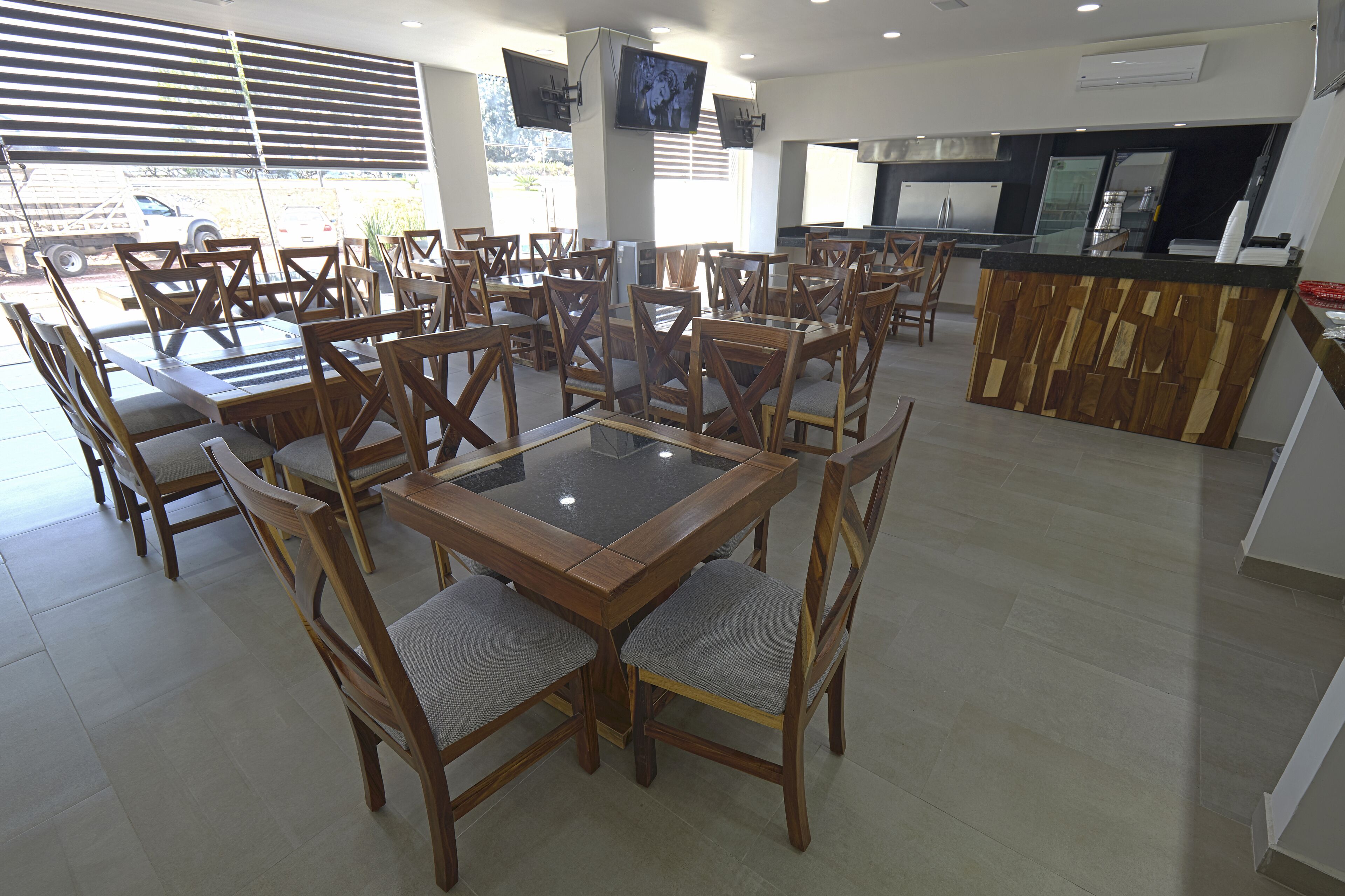 A spacious restaurant with multiple dining tables and chairs, a kitchen area, and large windows with blinds.