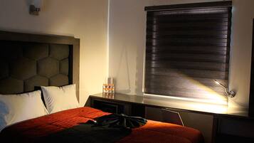 Quarto de hotel com cama, mesa de cabeceira, luminária e janela com persianas.