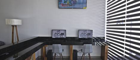 Un espace d’accueil d’hôtel avec un téléviseur à écran plat diffusant un programme, deux panneaux encadrés sur lesquels on peut lire « And Hotel », et un bureau moderne avec une lampe et des chaises.