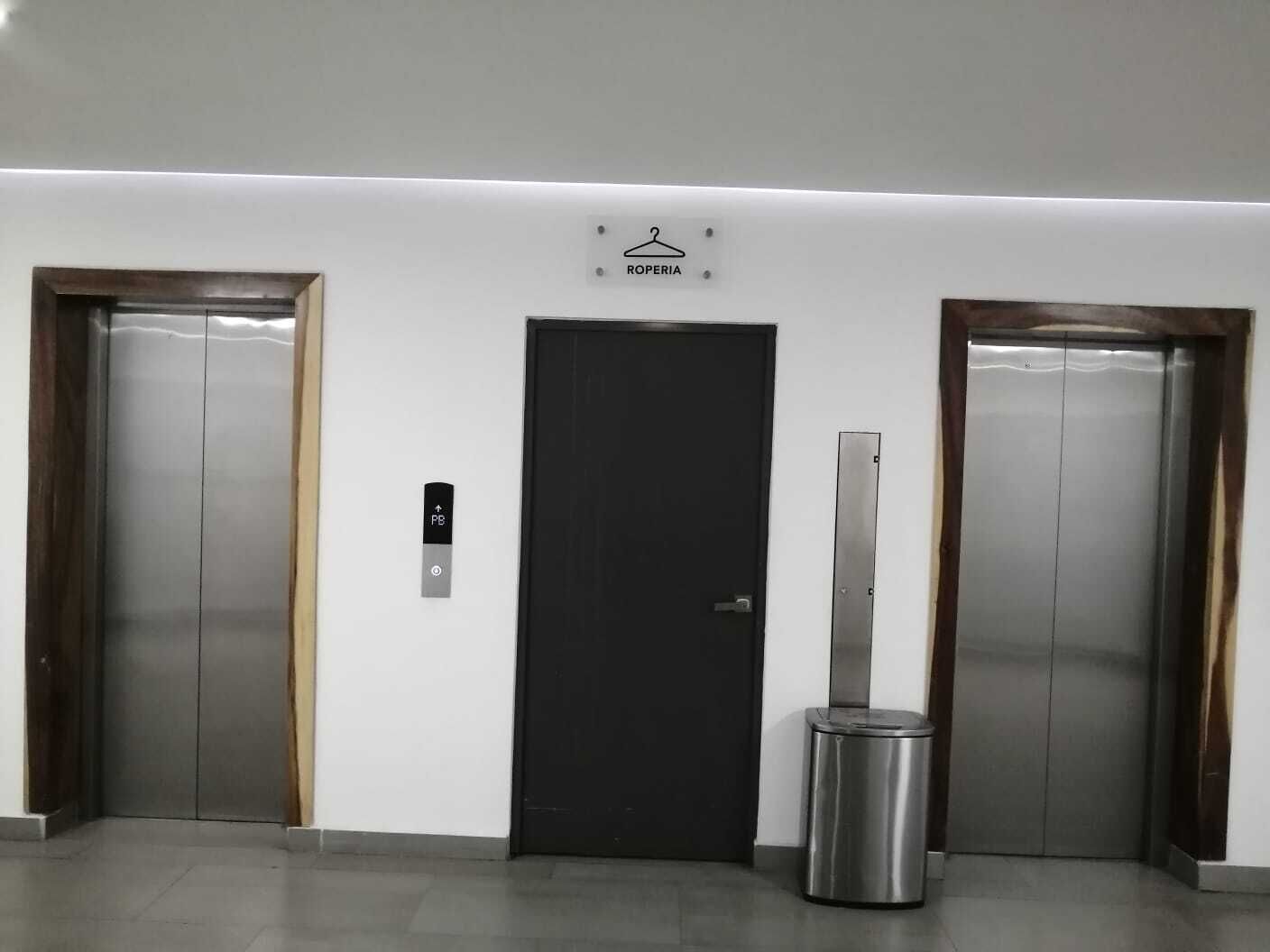 Un'area ascensori moderna, con due impianti di sollevamento, un cestino e una porta con il simbolo di un appendiabiti.