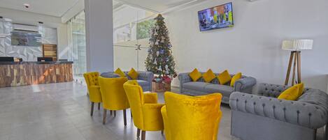 A modern lobby with a grey sofa, yellow chairs, a Christmas tree, and a TV on the wall.