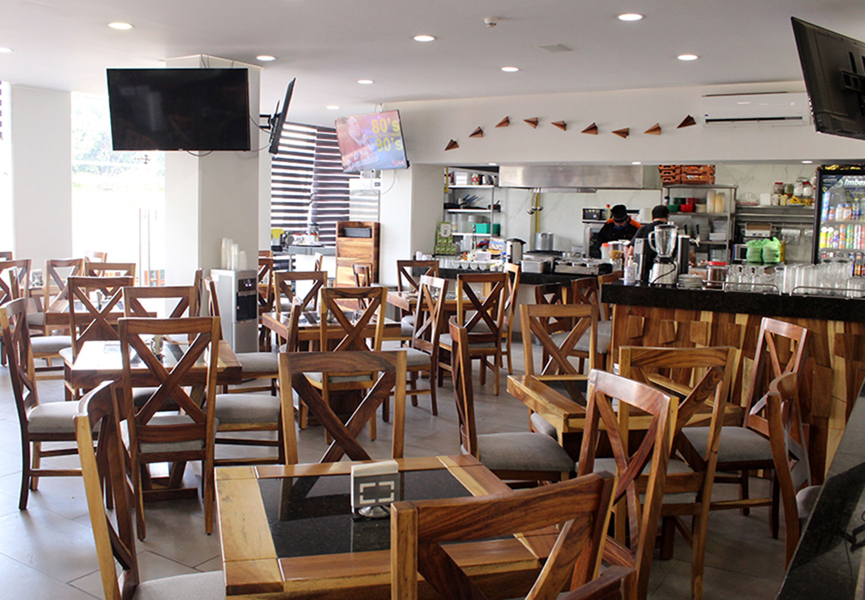 A restaurant with wooden chairs and tables, a kitchen area, and a flat-screen TV mounted on the wall.