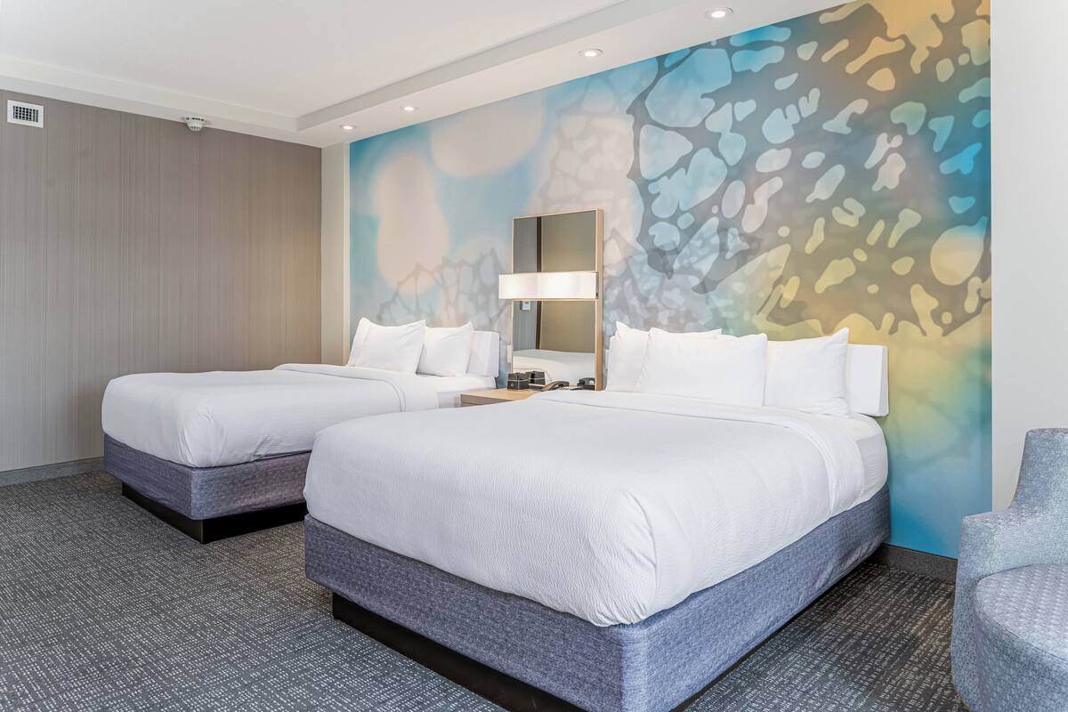 A hotel room with two beds, a large abstract mural, a modern lamp, and a gray armchair.