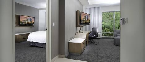 A hotel room with a bed, desk, chair, television, and a large window with a view of greenery.
