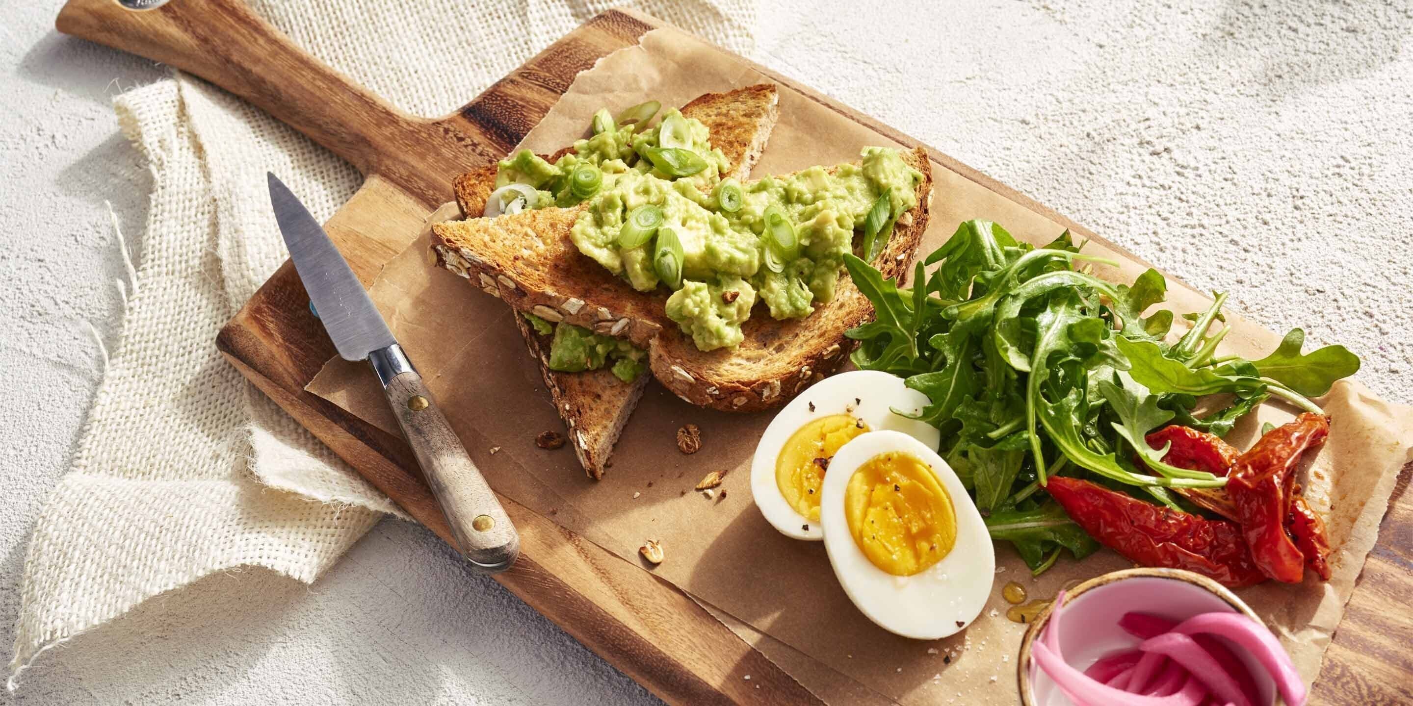A wooden cutting board with toast topped with avocado and a boiled egg, surrounded by greens and pickles.