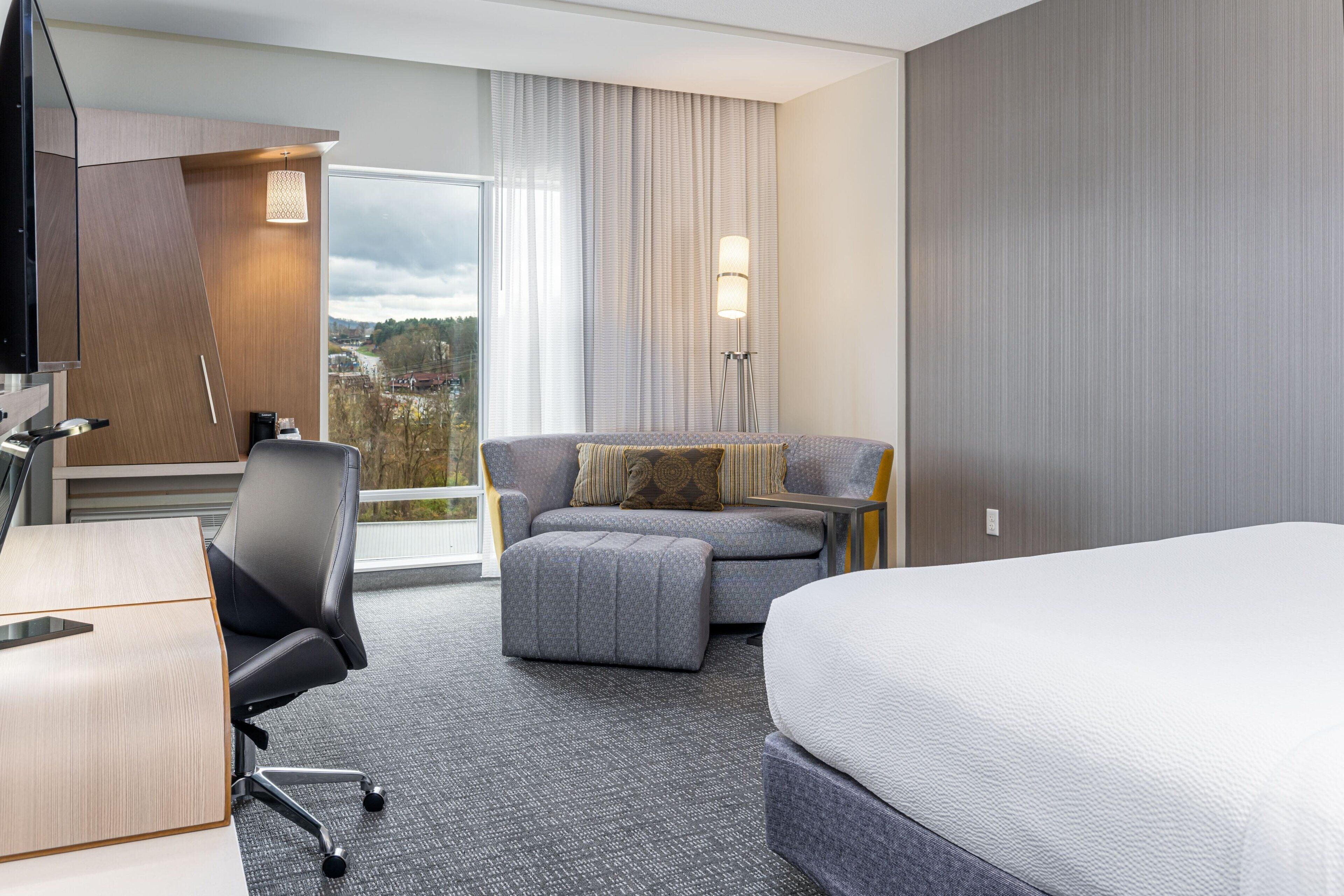 A hotel room with a bed, a desk, a chair, a sofa, and a view of the city.