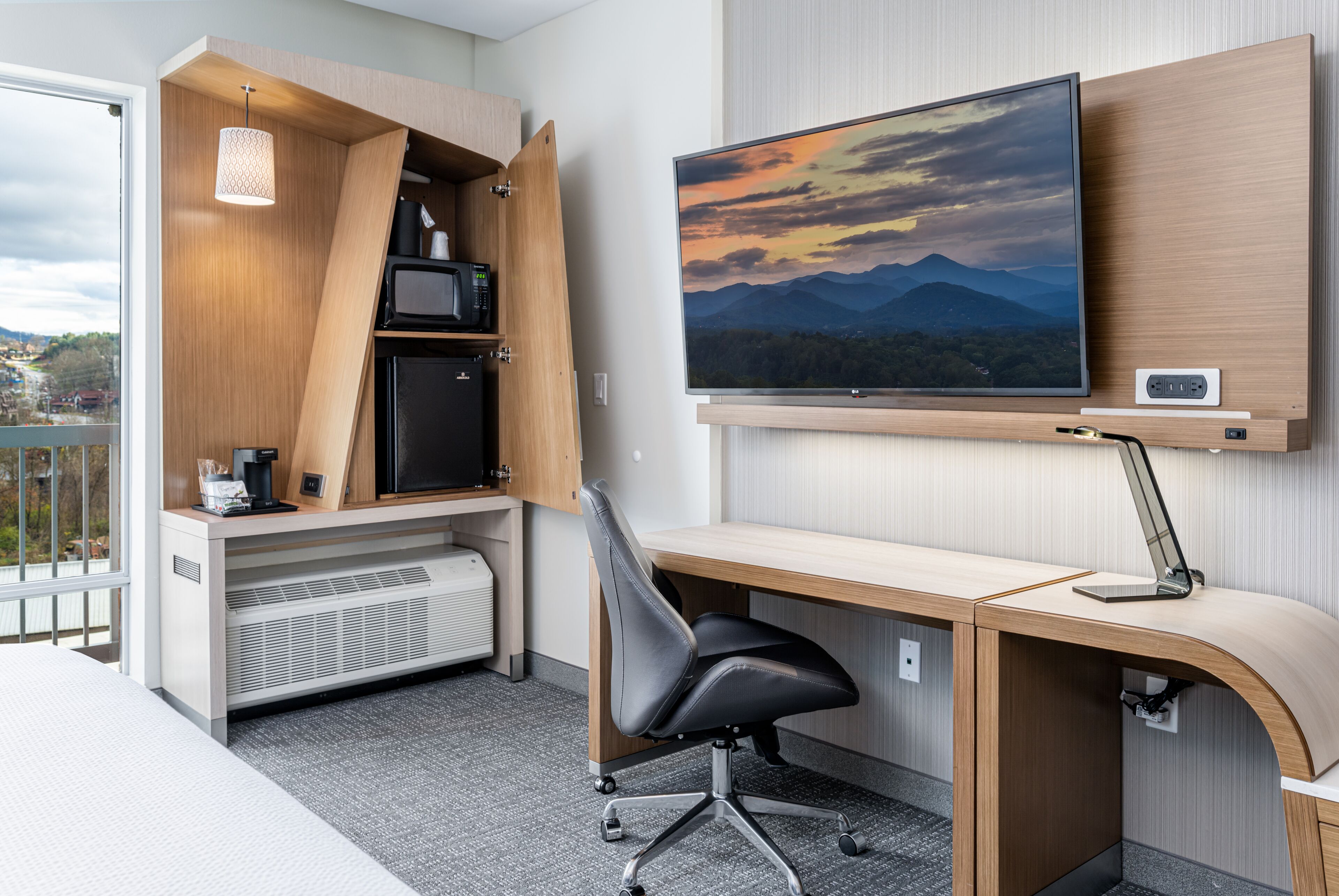 Quarto de hotel moderno com secretária embutida, televisão de tela plana, cadeira de escritório preta e micro-ondas.