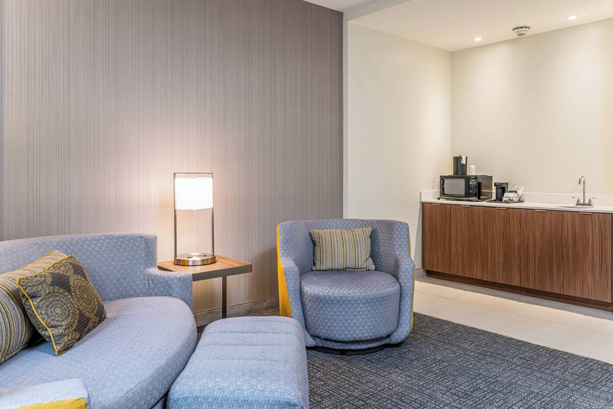 A modern hotel room with a grey sofa, a matching armchair, a small side table with a lamp, and a kitchenette area with a microwave and coffee maker.