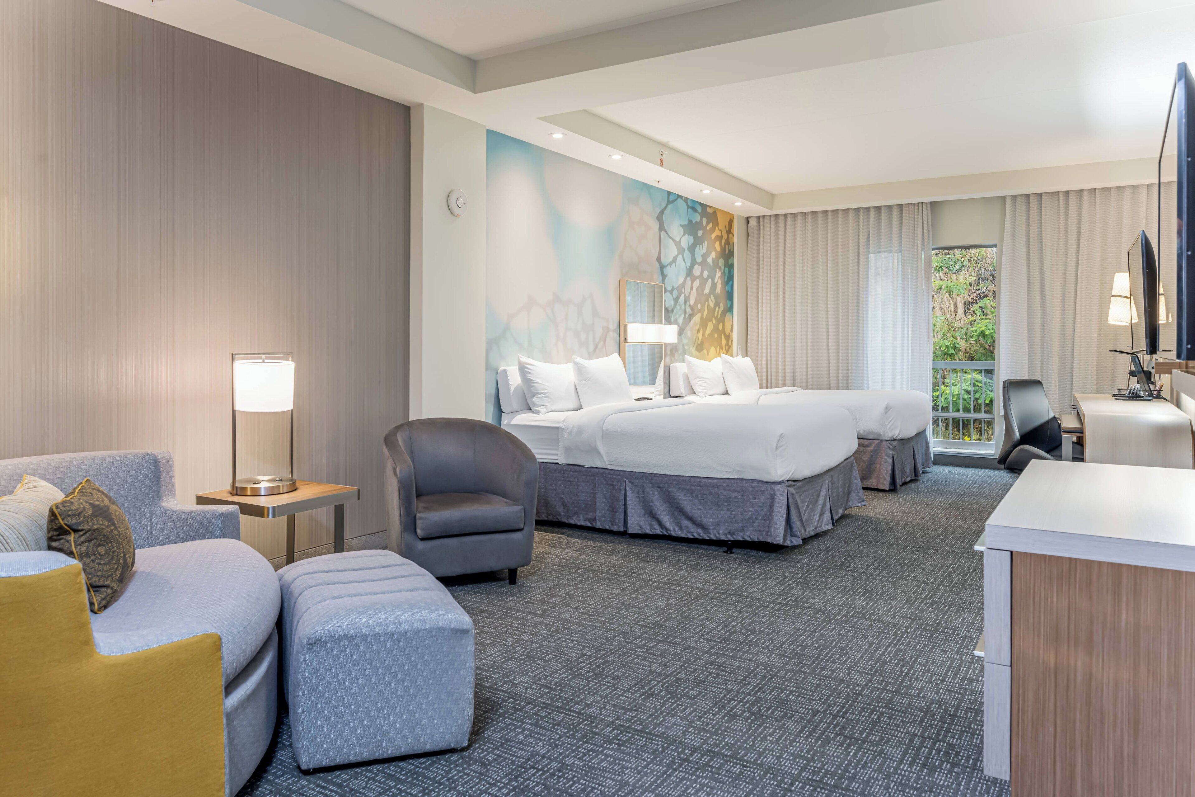Une chambre d’hôtel avec un grand lit, un canapé, un fauteuil, un bureau et une vue sur la verdure.