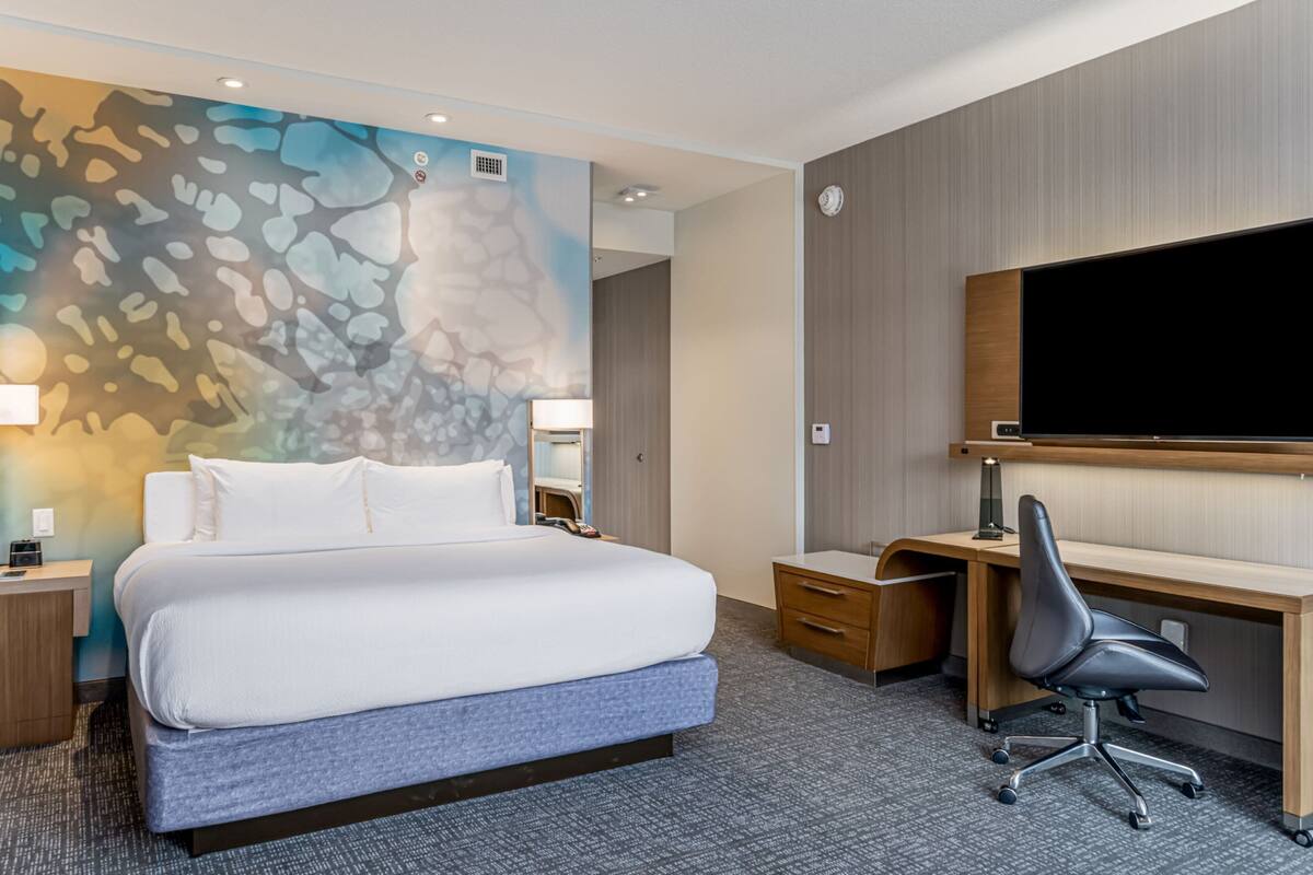 A hotel room with a large bed, a desk, a chair, a flat-screen TV, and a wall with a colorful abstract design.