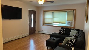 Una sala de estar con ventilador de techo, televisor de pantalla plana, ventana con persianas, sofá negro con cojines estampados y un pequeño reposapiés con control remoto.