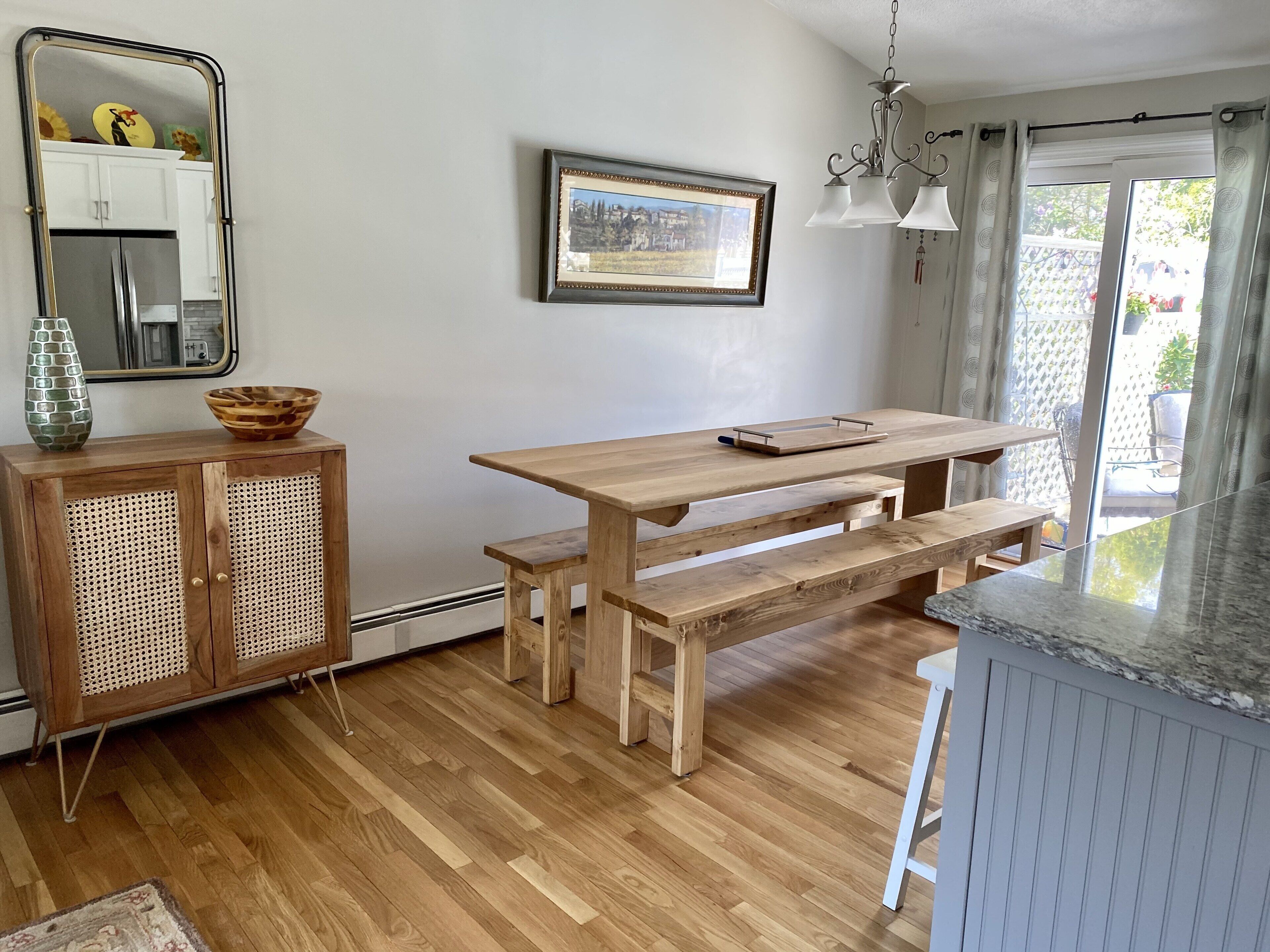 A kitchen with wooden cabinetry, a dining table with benches, a hanging light fixture, and a framed picture on the wall.