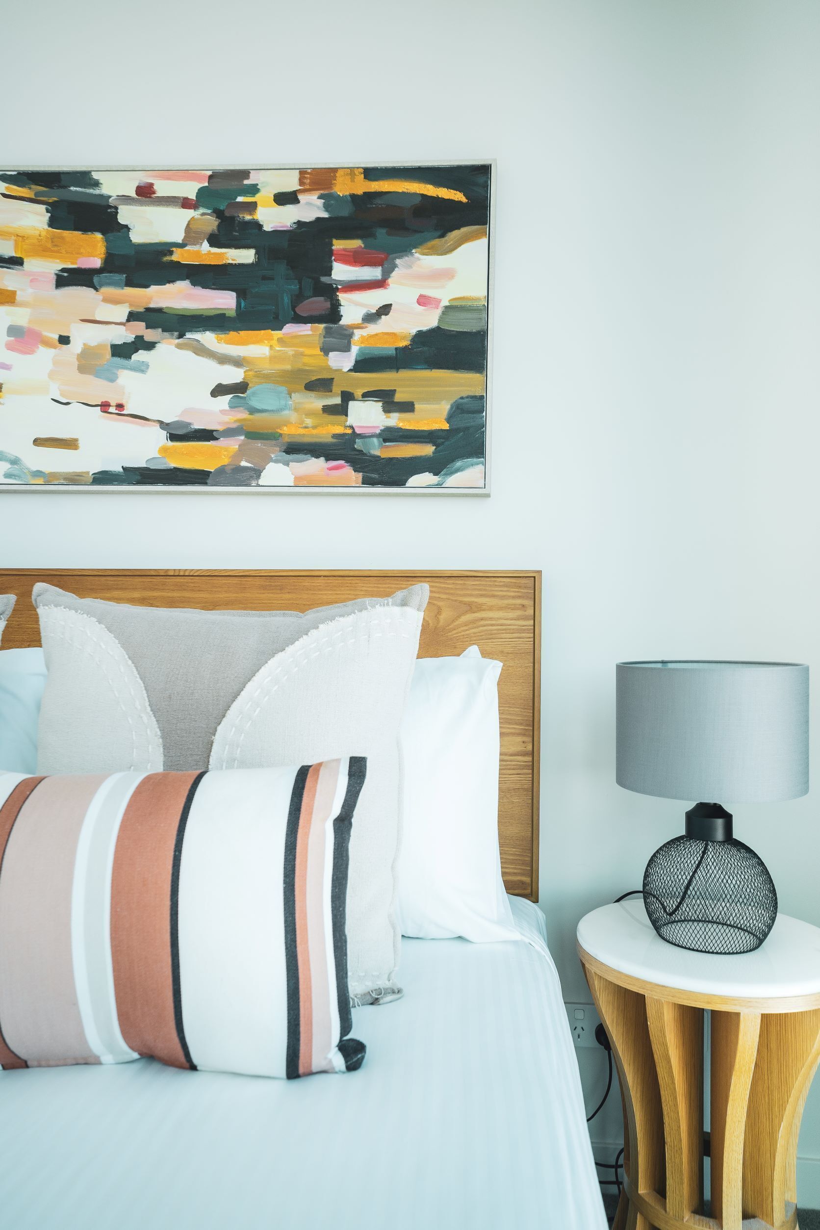 A neatly made bed with striped pillows, a round side table with a lamp, and a colorful abstract painting on the wall.