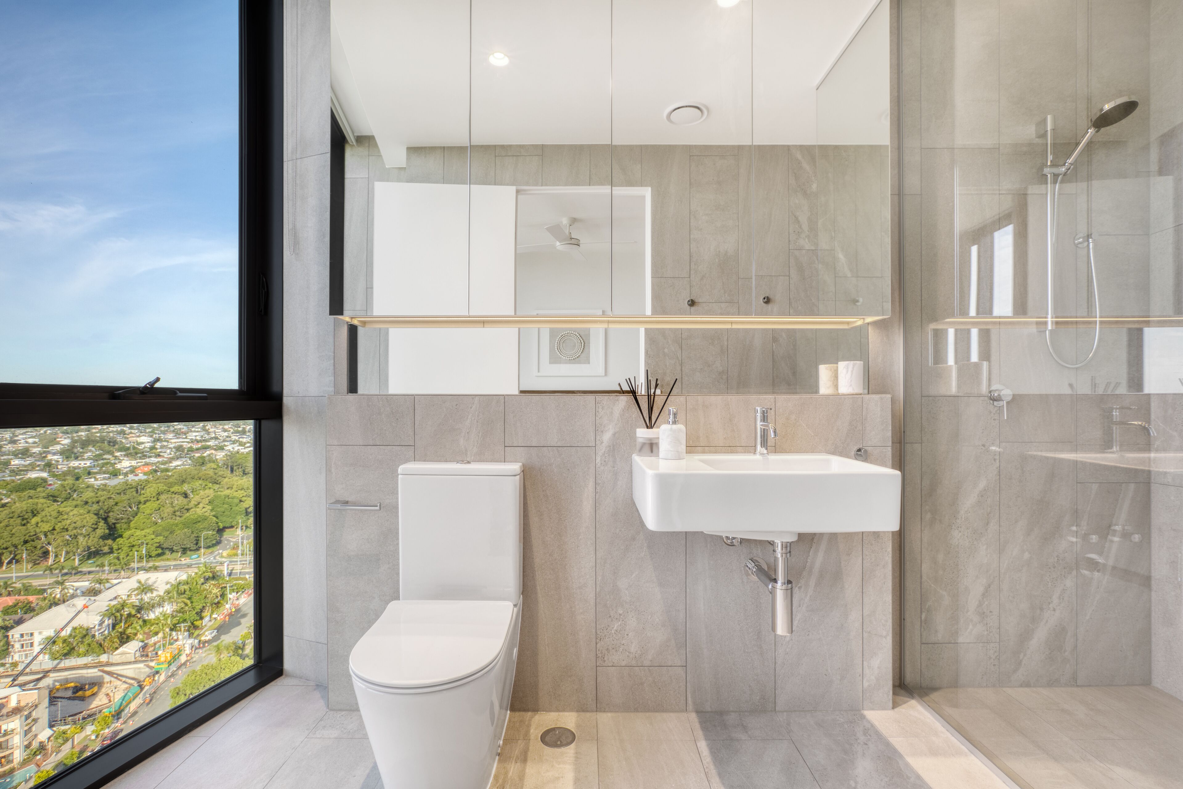 Ein modernes Badezimmer mit Toilette, Waschbecken und Dusche, großen Fenstern mit Blick auf die Stadt und einem minimalistischen Design.