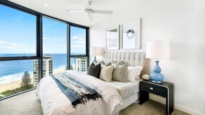 A bedroom with a large window overlooking a beach and high-rise buildings, a tufted headboard, and a bedside table with a lamp. - Vue Broadbeach (Broadbeach)