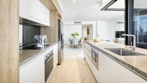 A modern kitchen with a central island, built-in appliances, and a large window offering a city view. - Vue Broadbeach (Broadbeach)