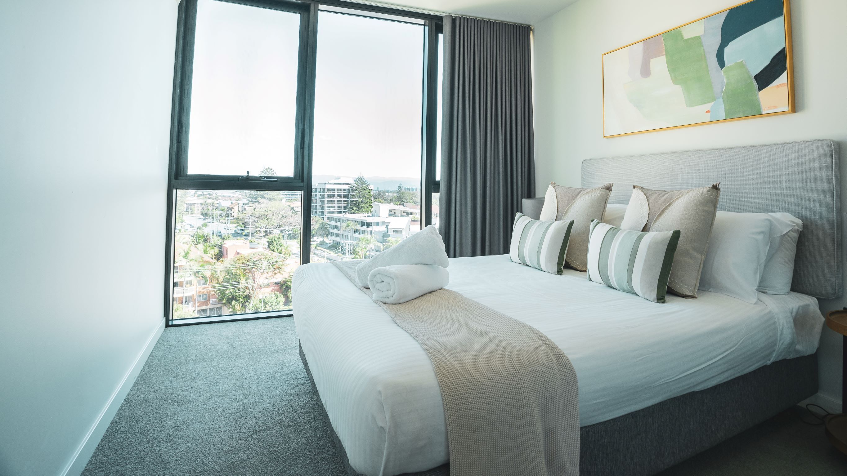 A hotel room with a large bed, a view of the city, and a painting on the wall.