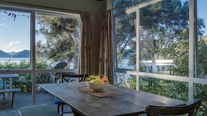 Un coin repas avec une table et des chaises en bois, un saladier rempli de fruits et de grandes fenêtres donnant sur un paysage de arbres et de montagnes.