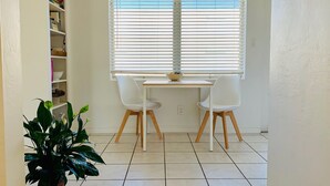 A room with a white tiled floor, a white desk with two chairs, a window with blinds, and a bookshelf with books and decorative items. -   Healthy, relaxing , romantic , organic vacation                   - (Naples)