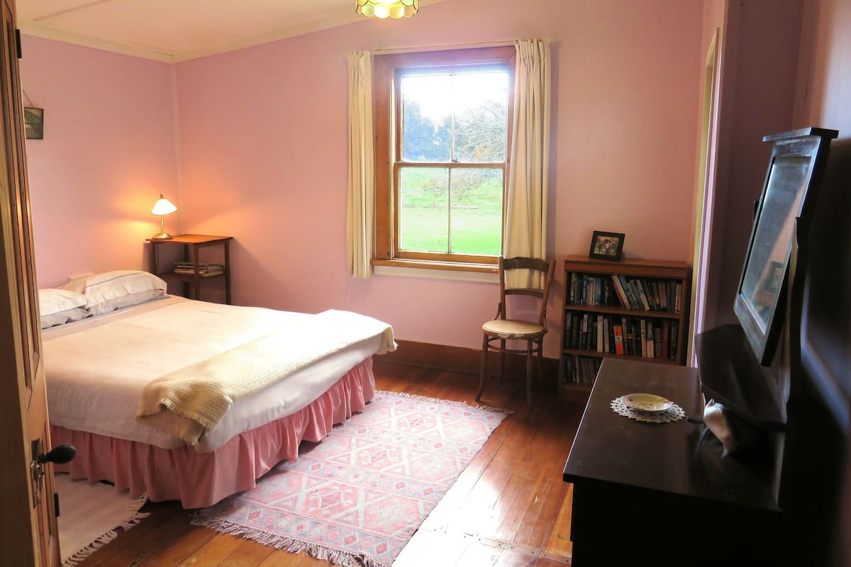 Une chambre à coucher avec un lit, une lampe de chevet, une fenêtre avec des rideaux, une chaise, une bibliothèque et un téléviseur posé sur une table en bois.