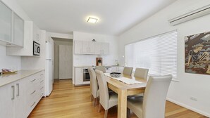 Une cuisine moderne avec des meubles blancs, un coin repas doté d’une table et de chaises en bois, et une grande fenêtre avec des stores.