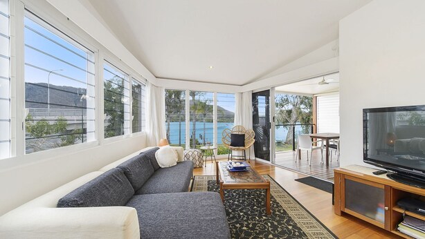 Un salon spacieux avec un grand canapé, une table basse et un téléviseur posé sur un meuble en bois. De grandes fenêtres offrent une vue sur la mer, et un balcon aménagé avec des sièges d’extérieur complète l’ensemble.