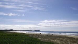 Une plage où l’on voit des gens se promener le long du rivage, un ciel dégagé et des collines à l’horizon.