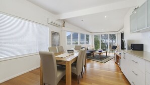 Une cuisine moderne avec des meubles blancs, un coin repas doté d’une table et de chaises en bois, et de grandes fenêtres offrant une vue sur l’extérieur.