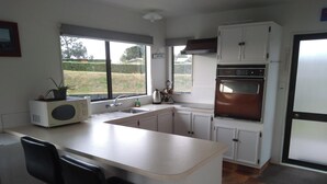 A kitchen with white cabinets, a built-in oven, a microwave, and a sink. There are two black bar stools and a window with blinds. - Arawai - Waimarama Holiday Home (Waimarama)