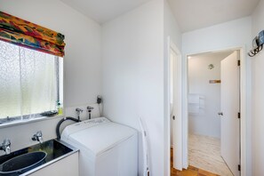Una habitación pequeña y de paredes blancas, con lavadora, lavabo y ventana con cortina de encaje.