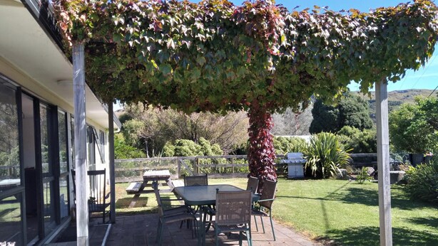 A covered patio with a table and chairs, a pergola with climbing plants, and a view of the surrounding garden and hills. - Arawai - Waimarama Holiday Home (Waimarama)