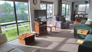 A spacious living room with a large window, a flat-screen TV, a grey sofa, a green sofa, and a wooden cabinet. - Arawai - Waimarama Holiday Home (Waimarama)