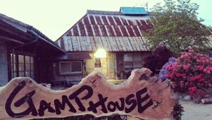 Un letrero de madera con la palabra "GAMPHOUSE" frente a un edificio rústico con techo de metal.