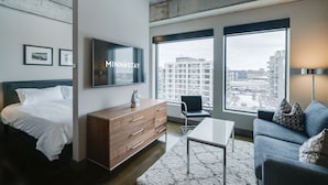 A modern hotel room with a large bed, a wooden dresser, a flat-screen TV, a sofa, and large windows offering city views.