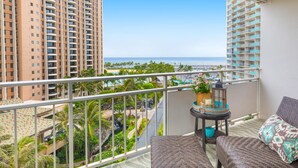 A balcony with wicker furniture, a small table with a lantern, and a view of a beach and palm trees. - Ilikai Luxury Suites (Honolulu)