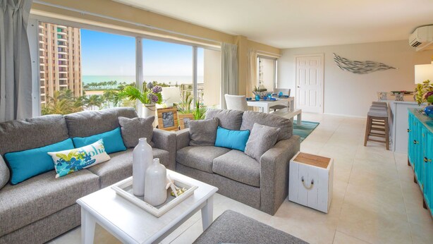 A modern living room with grey sofas, teal cushions, and a white coffee table. There is a dining area with a white table and chairs, and a kitchen area with teal cabinets. - Ilikai Luxury Suites (Honolulu)