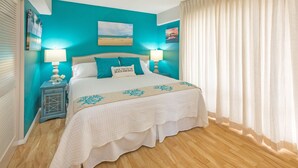 A bedroom with a bed, two bedside tables, a nightstand, a window with curtains, and a wall with a painting. - Ilikai Luxury Suites (Honolulu)