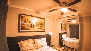 Ein Schlafzimmer mit einem Bett, einem Deckenventilator, einem Nachttisch, einer Lampe, einem Spiegel und einem Bild an der Wand.