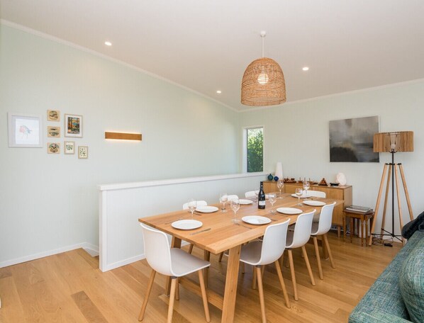 A dining area with a wooden table set for six, white chairs, a woven pendant light, and a floor lamp. - Ocean Oasis - Langs Beach Holiday Home (Langs Beach)
