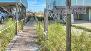 A wooden boardwalk leading to beach access, flanked by buildings with a sign reading "Summer House West Beach Access".