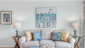 A living room with a light-colored sofa, two side tables with lamps, a blue coffee table, and a painting of seagulls on a pier.