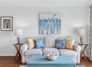 A living room with a light-colored sofa, two side tables with lamps, a blue coffee table, and a painting of seagulls on a pier. - Perfect Couples Getaway ^ Fun, Sunsets, Beach Walks ^ Quiet End of West Beach (Gulf Shores)