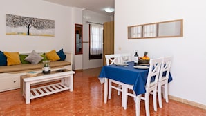 A dining area with a table set for four, white chairs, and a blue tablecloth. There is a white sofa with colorful cushions in the background, a small white table with a glass jar and a book, and a painting on the wall. - Apartment Lima Center Playa Blanca By PVL (playa blanca)