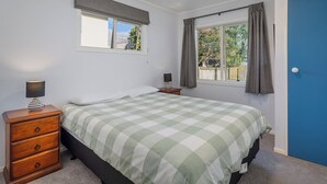 A bedroom with a bed, a dresser, a window with curtains, and a door. - Nest by the Beach - Cooks Beach Holiday Home (Cooks Beach)