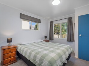 A bedroom with a bed, a dresser, a window with curtains, and a door. - Nest by the Beach - Cooks Beach Holiday Home (Cooks Beach)