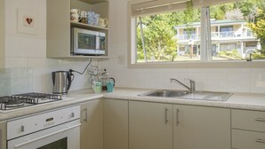 Une cuisine avec des meubles blancs, un évier en acier inoxydable, un four à micro-ondes, une cuisinière et une fenêtre donnant sur un immeuble et des arbres.