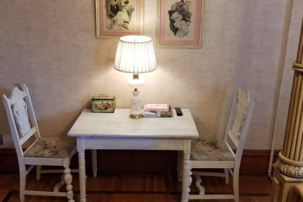 A white table with a lamp, a vintage radio, and books. Two chairs with floral upholstery are positioned on either side of the table.