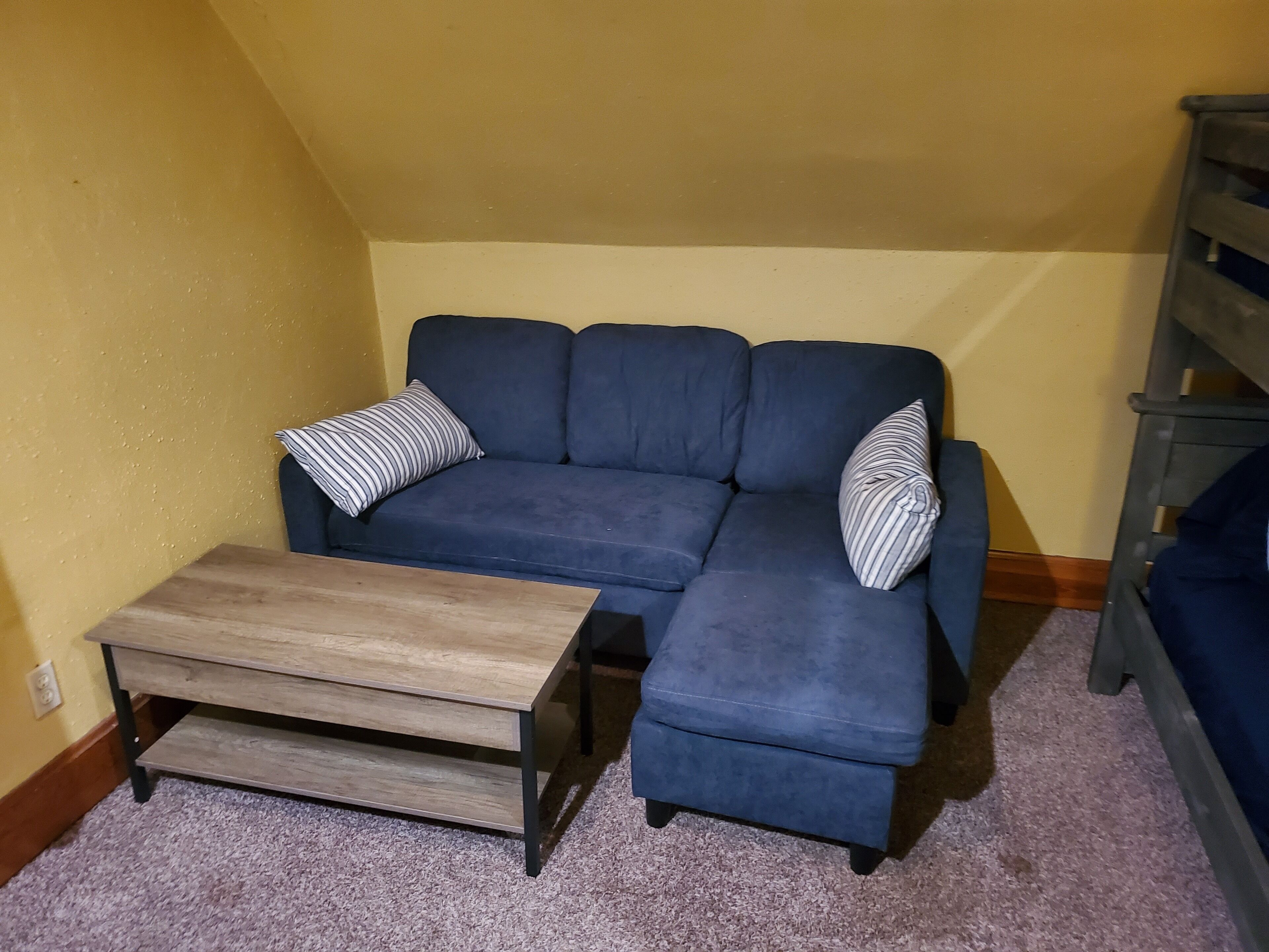 A blue sectional sofa with striped pillows, a wooden coffee table, and a carpeted floor in a room with yellow walls and a wooden bench in the corner.