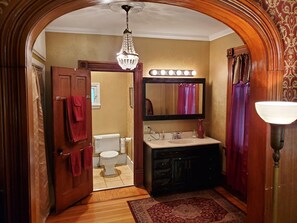A bathroom with a wooden door, a chandelier, a vanity with a mirror, and a red rug.